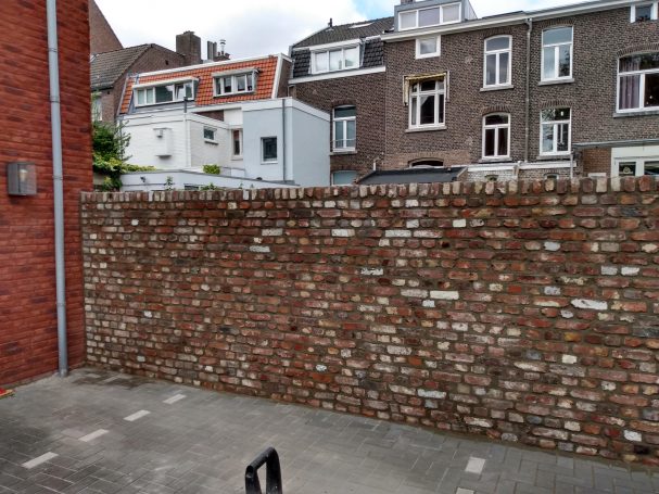 Mauer aus roten Ziegeln, dahinter schematische Gebäudeansichten.